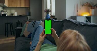 A woman uses a mobile phone with green screen while lying on the couch at home. A rear view of her hands, an unrecognizable person - person photo