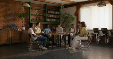 A group of young people are smiling and talking while sitting around a round table indoors. Notebooks and books on the table. Daylight - sitting photo