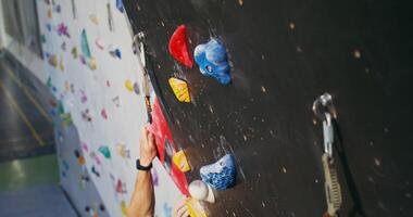 A young man, with a focused look, climbs up the climbing wall, attaching a safety rope to the hooks on the wall. Indoor gym overhead filming with artificial lighting - attaching photo