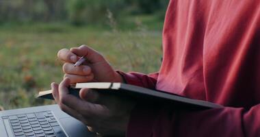 A young man of European appearance makes notes in a notebook, using a laptop, sitting on the ground under a tree, close-up - sitting photo