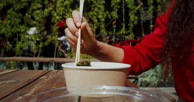 A young woman eats Asian food out of a box using chopsticks while sitting at a table on the cafe terrace, hands close-up, no face - chopsticks photo
