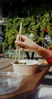 A young woman in casual clothes eats Asian food out of a box using chopsticks while sitting at a table on the cafe terrace, hands close-up, no face - chopsticks photo