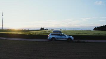 A car with a roof rack drives along a road through picturesque green meadows and arable fields on a beautiful sunny morning. Wind generator in the rays of the rising sun - picturesque photo