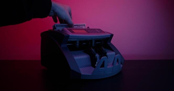 A man's hand inserts a stack of hundred dollar bills into a counting machine and it starts counting, studio photo