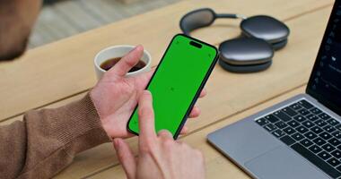 The man uses a mobile phone with green screen. He is sitting in front of an open laptop at a table. An unrecognizable person photo