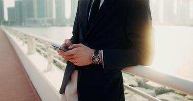 A man in a business suit with a wristwatch on his hand uses a smartphone standing on the embankment on a sunny day. Men's hands close-up - smartphone photo
