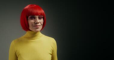 A woman with glasses and an orange wig smiles at the camera, holding a mobile phone with green screen in her hand, standing on a gray background in the studio photo