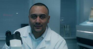 A man in a lab coat looks at the camera sitting in front of a microscope in a medical laboratory photo