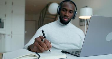 A man with headphones is talking on a chat on a laptop, sitting at a table at home. He writes in a notebook. Remote work - Remote photo