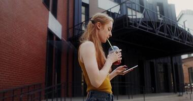 A young red-haired woman uses a mobile phone and drinks juice from a disposable cup with a straw while walking on a city street photo