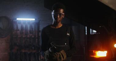 A young blacksmith of African American appearance in goggles and an apron turns on an angle grinder and begins to cut off a piece of metal workpiece while working in a forge. 4k, red komodo - American photo