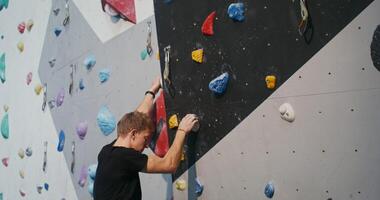A young man of European appearance, wearing a safety belt, is concentratedly climbing up the climbing wall, grabbing the artificial ledges imitating stones photo