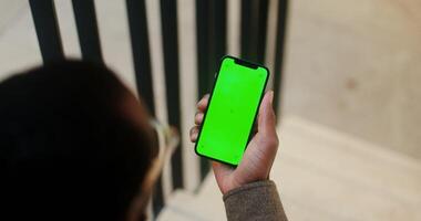 An African American man holds a mobile phone with a green screen in his hand. filming from behind, his face is not visible. Copy space, mockup, chroma key. in 4k, red komodo - mobile photo