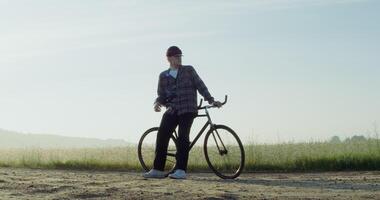A young man with a camera around his neck stopped near a wheat field, resting after cycling and admiring the beauty of nature in the rays of the rising sun. 4k , red komodo - stopped photo