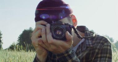 A young male photographer adjusts his camera and takes a picture while standing among wheat ears in an agricultural field on a sunny day, close-up. 4k , red komodo - camera photo