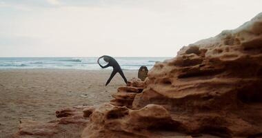 A man in a diving suit does exercises standing on the sandy seashore next to a surfboard stuck in the sand. Rear view - standing photo