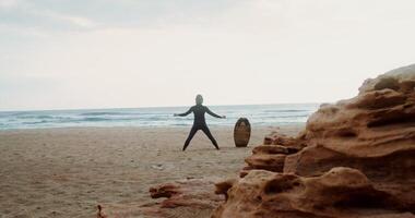 A man in a diving suit does exercises standing on the sandy seashore next to a surfboard stuck in the sand. Rear view photo