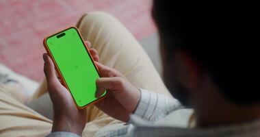 A man in casual clothes scroll the green screen of a mobile phone while sitting on the couch at home. Top view, no face, close-up of a man's hands - scroll photo