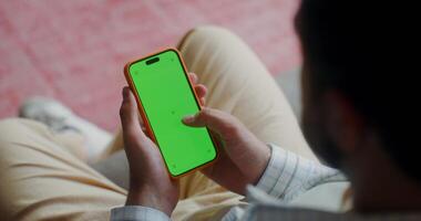 A man in casual clothes scroll the green screen of a mobile phone while sitting on the couch at home. Top view, no face, close-up of a man's hands photo