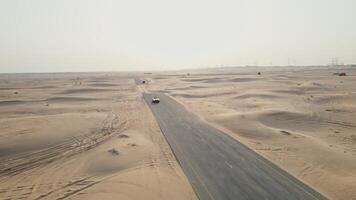 A yellow car is driving along an asphalt road in the desert, raising sand dust behind it. Drone photo
