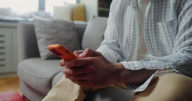 A man in casual clothes smiles while typing on a mobile phone while sitting on the couch at home. The moves from the man's hands to his face photo