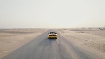 Rear view of a yellow car driving along the road among the desert sands on a sunny day. Drone view photo