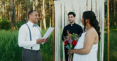 The groom reads the oath at the wedding ceremony, standing with the bride at the altar in front of the priest outdoors - wedding photo