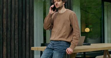 A man in casual clothes is talking on a mobile phone standing on the terrace of a stylish cottage on a summer day. An open laptop is on the table photo