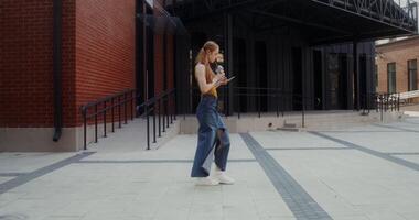 A young red-haired woman uses a mobile phone and drinks juice from a disposable cup with a straw while walking on a city street - walking photo