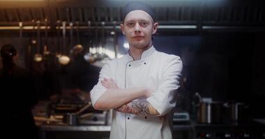 A man in a chef's uniform with a headband looks at the camera with a slight smile, standing in a modern kitchen with his arms crossed at his chest - camera photo