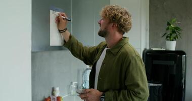A man in casual clothes fills out a chart with a schedule hanging on the kitchen cabinet. Daylight, home interior - hanging photo