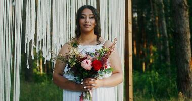 An African-American bride with a bouquet in her hands stands at a decorated arch in the forest and smiles looking at the camera photo