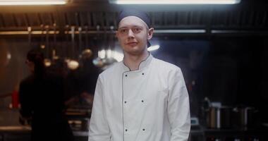 A man in a chef's uniform with a headband looks at the camera with a slight smile, standing in a modern kitchen with his arms crossed at his chest - slight photo