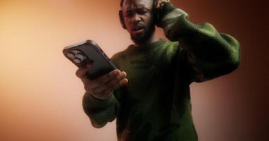 A bearded man in a sweater and headphones turns on music on his mobile phone and starts dancing standing on an orange background in the studio photo