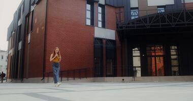 A young red-haired woman uses a mobile phone and drinks juice from a disposable cup with a straw while walking on a city street - disposable photo