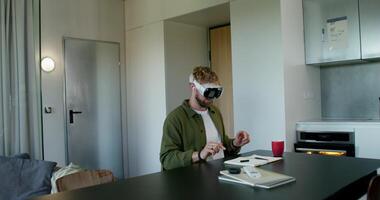A man in a virtual reality headset typing on a virtual keyboard sitting at a table at home. Daylight photo