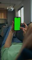 A woman uses a mobile phone with green screen lying on the couch at home. A rear view of her hands, an unrecognizable person - unrecognizable photo