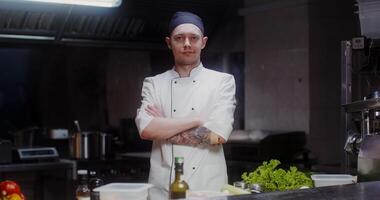 A man in a chef's uniform with a headband looks at the camera with a slight smile, standing in a modern kitchen with his arms crossed at his chest photo