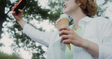 A young woman takes a selfie on a mobile phone standing in a park with ice cream in her hand photo