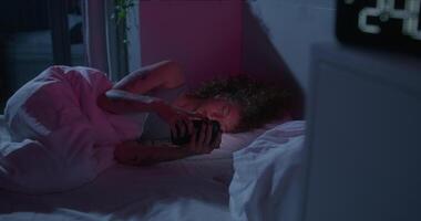 A woman uses a mobile phone lying on the bed. Dark room, home interior. The electronic clock is on the bedside table photo