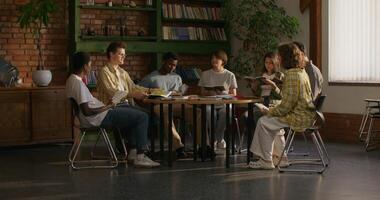 A group of young people of different nationalities join hands and begin prayer while sitting at a table indoors. Daylight photo