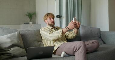 A bearded man in casual clothes is talking on a chat on a mobile phone sitting on the couch at home. The laptop is next to him. Daylight photo