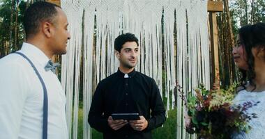 A young priest with a tablet in his hands marries an African-American couple in a beautiful pine forest near a decorated arch - couple photo