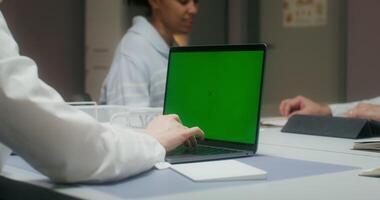 The nurse uses a laptop with a green screen, entering patient data. A woman talks to a doctor sitting at a table in the background - patient photo