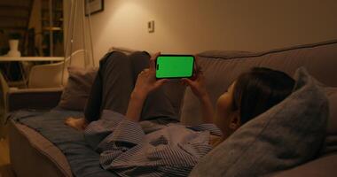 A woman uses a mobile phone with a green screen lying on the couch at home, dimmed evening light - mobile photo
