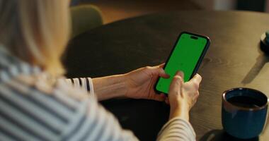 A woman uses a mobile phone with green screen sitting at a table indoors, close-up of her hands, an unrecognizable person - unrecognizable photo