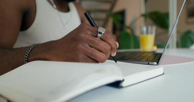 A man uses a laptop and writes in a notebook sitting at a table at home, close-up of his hand, an unrecognizable person photo