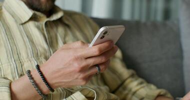 A man is typing on a mobile phone sitting on the couch at home. close-up of his hands, an unrecognizable person. Daylight - unrecognizable photo