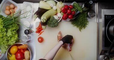 A woman in disposable black gloves cuts bacon while working at a table full of vegetables, close-up of her hands, top view photo