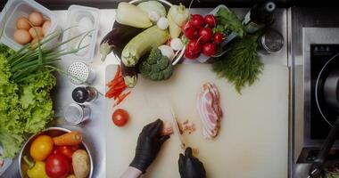 A woman in disposable black gloves cuts bacon while working at a table full of vegetables, close-up of her hands, top view - working photo